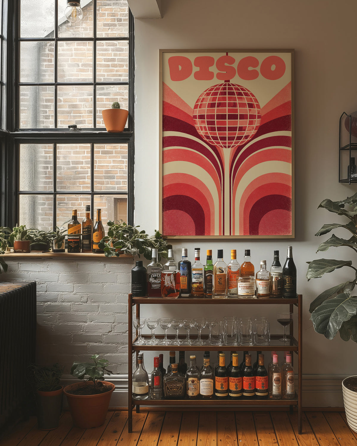 Rose Mirror Ball After Midnight, a retro disco ball drawn in a grid of rose and cream lines at the center of stacked arches in rose, cream and oxblood, with the word DISCO in bold rose letters across the top, in a wood frame on a brick wall above a bar cart of bottles and plants