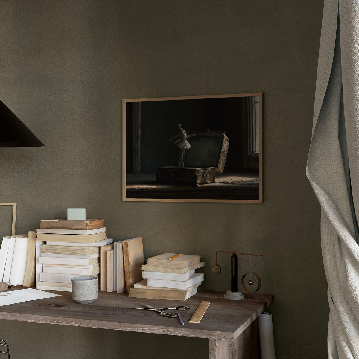 She Waits Before the First Note, a porcelain ballerina figurine poised on a carved music box in a dim room, painted in charcoal, slate and pale ivory, in a thin wood frame on a moss green wall above a desk stacked with books
