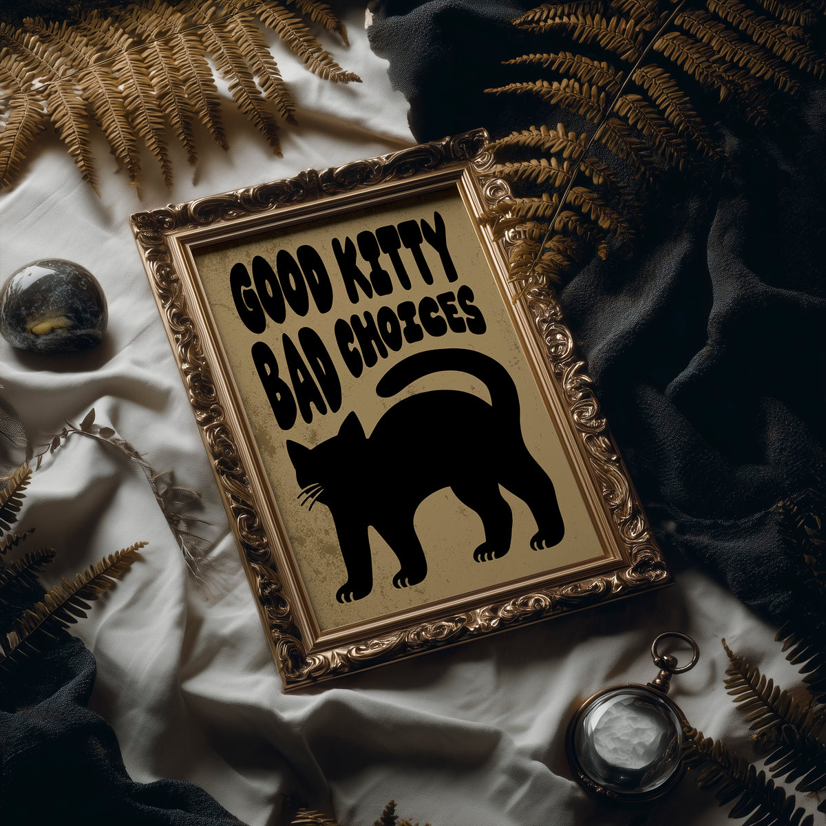 Sleek Silhouette, Questionable Taste, a solid black cat silhouette with a long tail curled up the side, standing on tan paper with the words GOOD KITTY BAD CHOICES in bold black lettering, in an ornate frame on silk cloth beside ferns and a brass compass