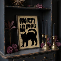 Sleek Silhouette, Questionable Taste, a solid black cat silhouette with a long tail curled up the side, standing on tan paper with the words GOOD KITTY BAD CHOICES in bold black lettering, in a dark frame on a black shelf beside brass candles and dried leaves