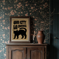Sleek Silhouette, Questionable Taste, a solid black cat silhouette with a long tail curled up the side, standing on tan paper with the words GOOD KITTY BAD CHOICES in bold black lettering, in a dark wood frame on a floral wallpaper wall above a rustic cabinet and a vase