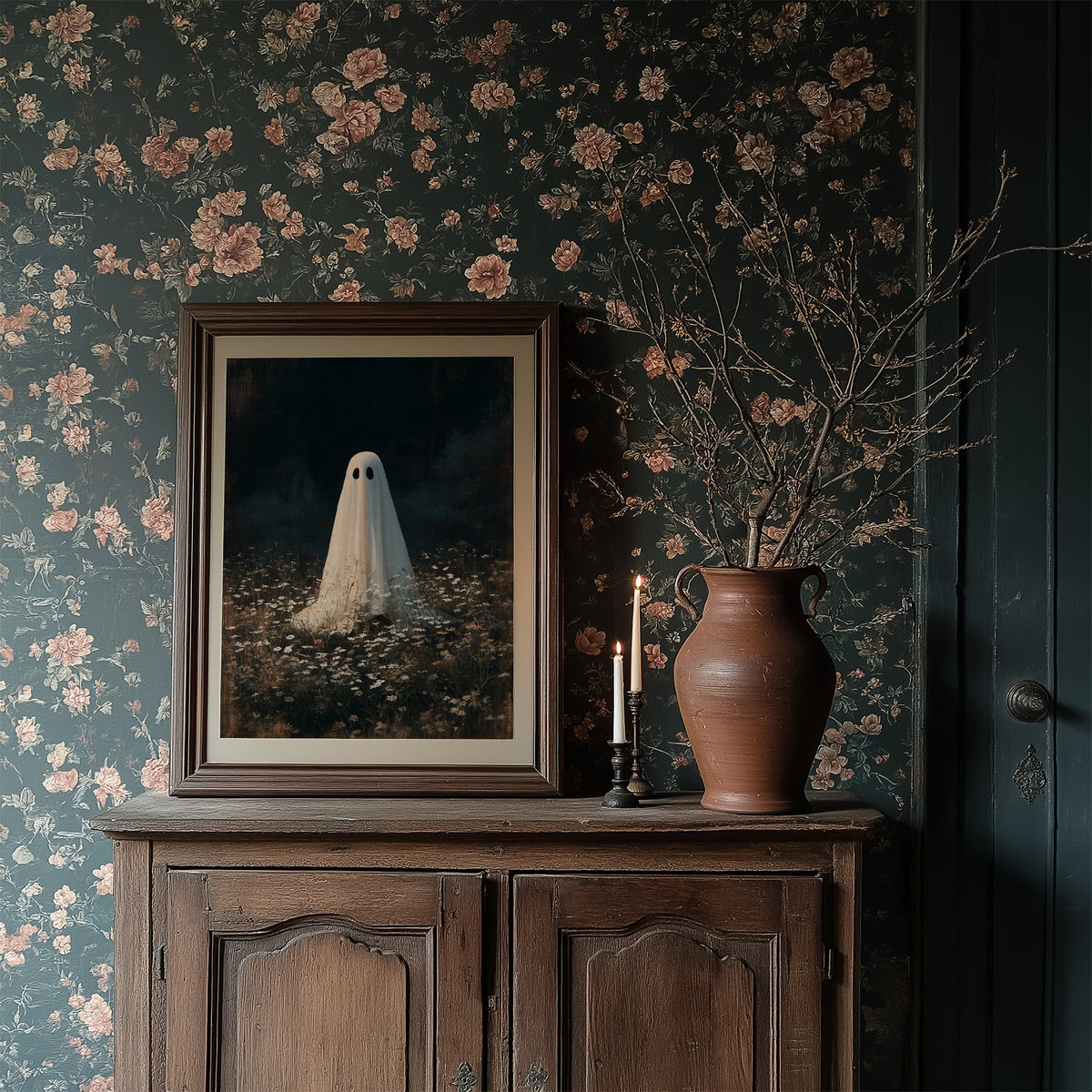 Someone Stayed Behind in the Meadow, a small white ghost standing in a dark field of pale wildflowers beneath a black woodland, painted in deep green, charcoal and bone, in a dark wood frame on a floral wallpaper wall above a rustic cabinet and a vase
