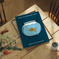 Still Enough for Today, a round glass fishbowl with one goldfish on a deep navy ground, a line of cream lettering above and below in a cartoon style, painted in navy, pale blue and orange, lying flat on a light wood table beside a coffee cup and red berries