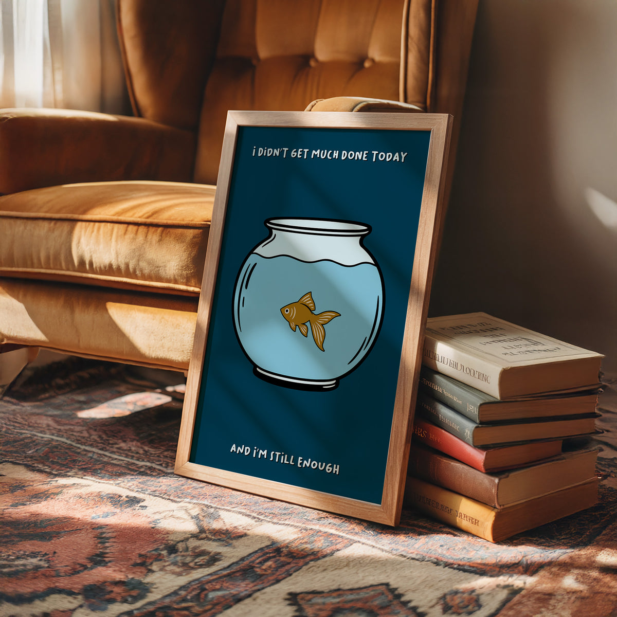Still Enough for Today, a round glass fishbowl with one goldfish on a deep navy ground, a line of cream lettering above and below in a cartoon style, painted in navy, pale blue and orange, in a wood frame on a rug beside a leather armchair and a stack of books