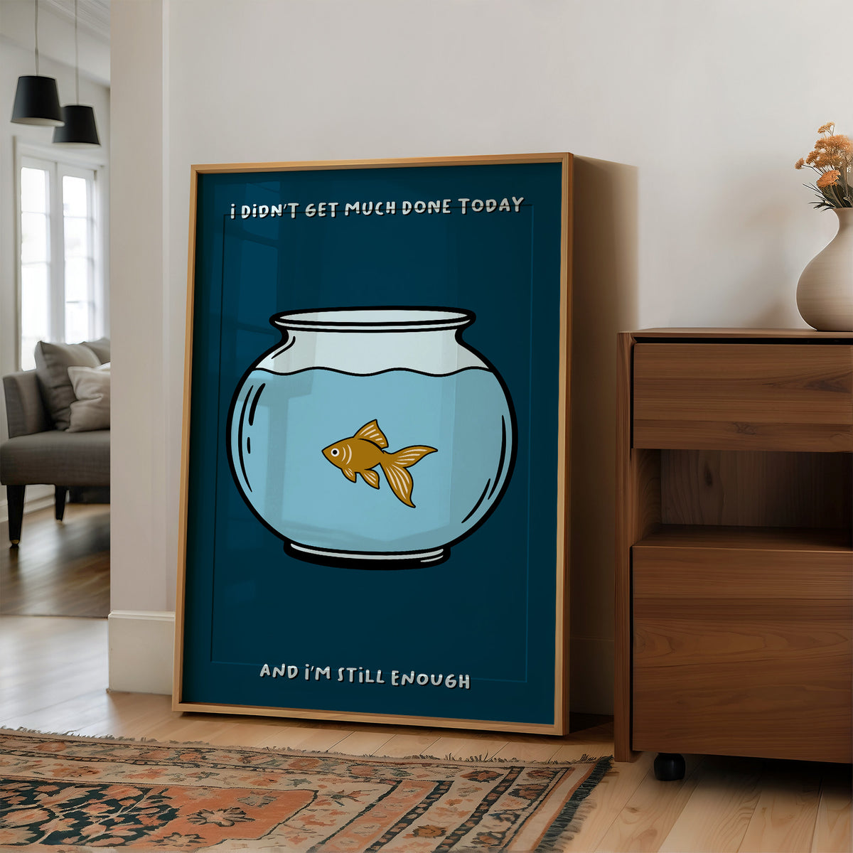 Still Enough for Today, a round glass fishbowl with one goldfish on a deep navy ground, a line of cream lettering above and below in a cartoon style, painted in navy, pale blue and orange, in a wood frame on a wall beside a walnut cabinet and an open doorway
