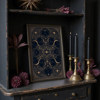 The Gilded Wheel of Midnight, a gold ornamental mandala of moon phases and stars on a deep navy field, painted in navy and antique gold, on a dark wooden cabinet shelf with candles and dried flowers