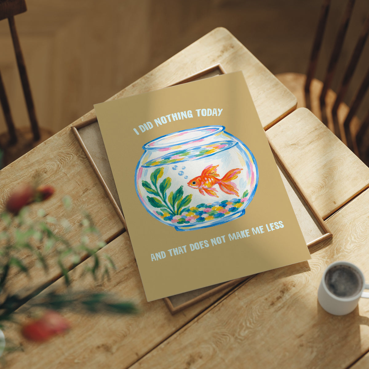 The Goldfish Takes the Afternoon, a goldfish circling a round bowl of pale blue water with green plants, with three lines of cream lettering on a mustard ground, in a flat playful illustrated style painted in mustard, blue and cream, lying flat on a light wood table beside a coffee cup and red berries