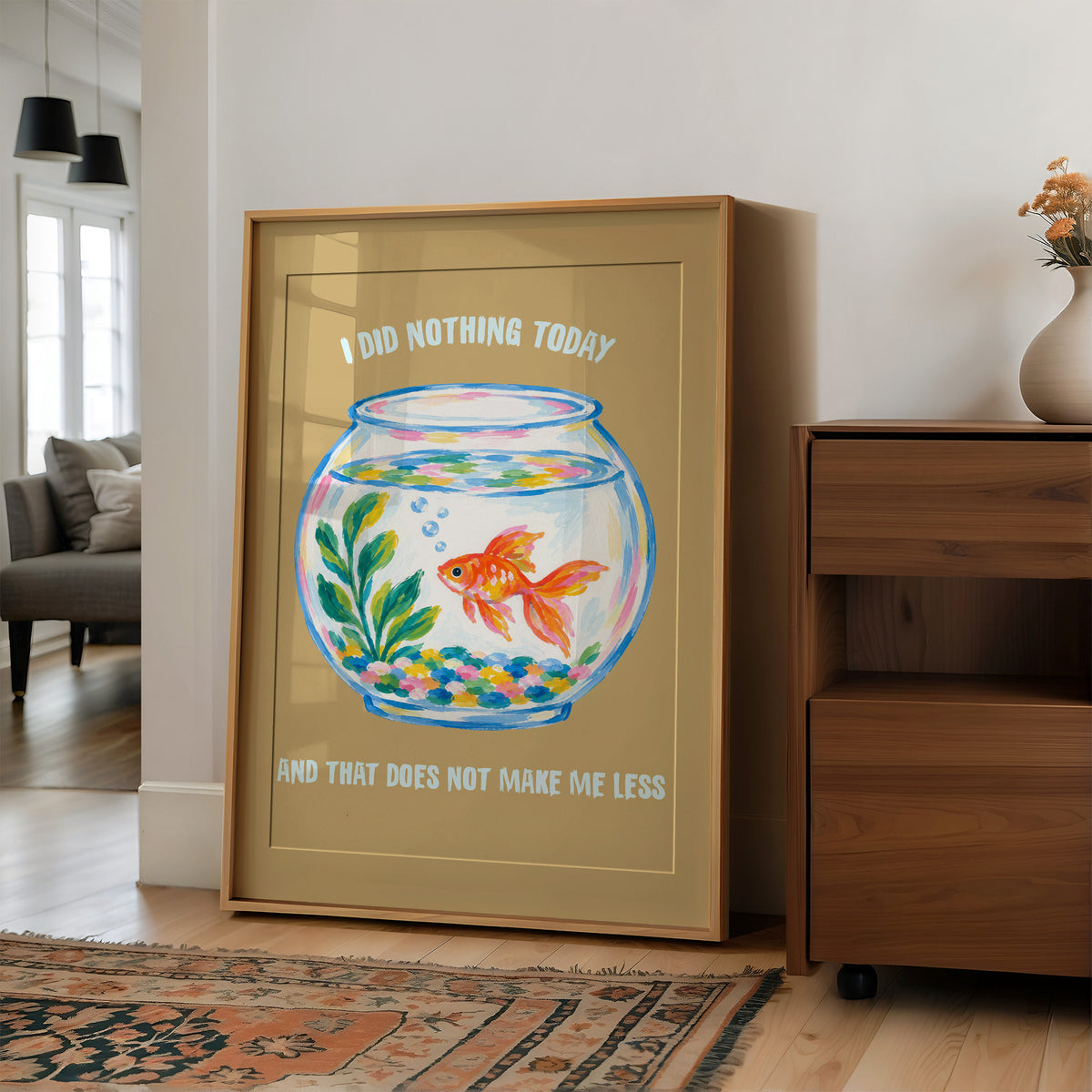 The Goldfish Takes the Afternoon, a goldfish circling a round bowl of pale blue water with green plants, with three lines of cream lettering on a mustard ground, in a flat playful illustrated style painted in mustard, blue and cream, in a wood frame on a wall beside a walnut cabinet and an open doorway