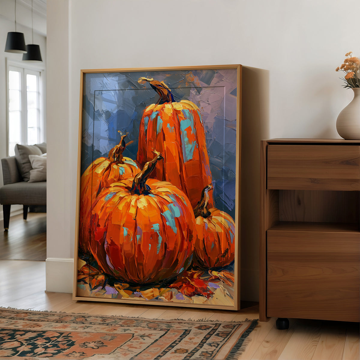The Harvest Keeps Its Blue Hour, three painted pumpkins in orange and teal on a dusky blue ground with fallen leaves at their feet, in a loose, bold, painterly style, in a wood frame on a wall beside a walnut cabinet and an open doorway