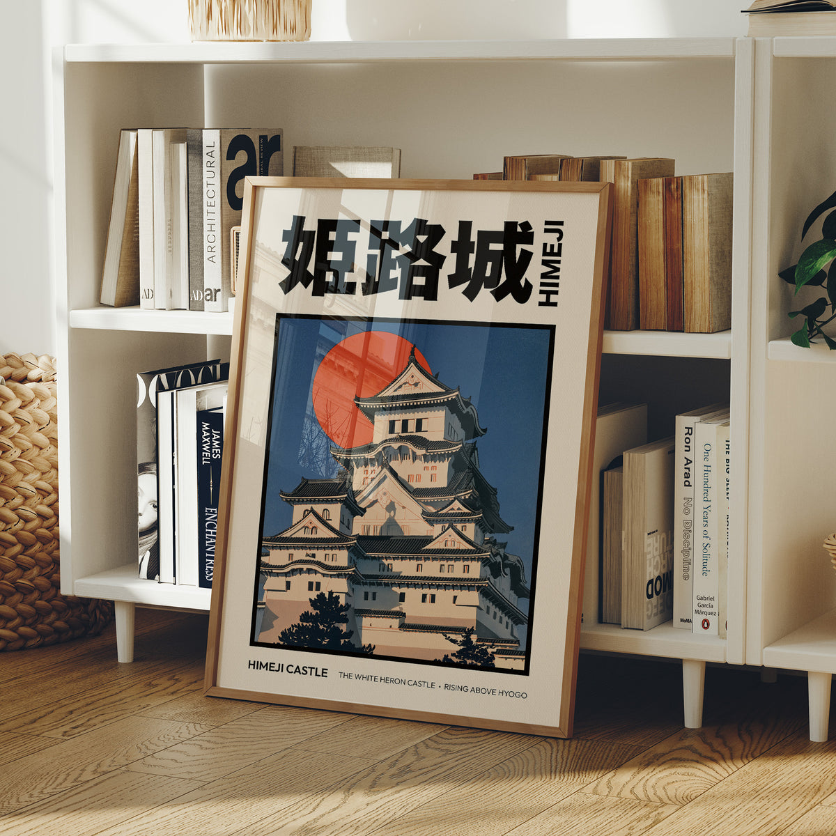 The Hill Keeps Its Pale Towers, a hilltop castle with tiered black roofs and white walls above a rising vermilion sun, in a flat Japanese travel poster style with bold black lettering on cream, painted in indigo, cream and vermilion, in a wood frame on a white bookshelf beside stacked books and a small basket