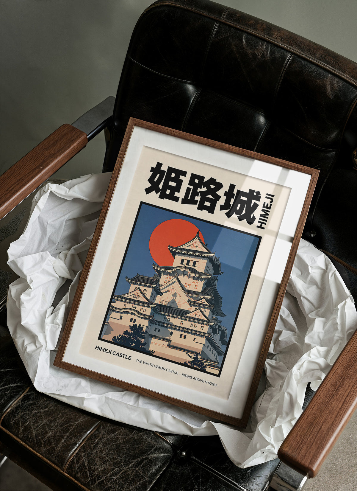 The Hill Keeps Its Pale Towers, a hilltop castle with tiered black roofs and white walls above a rising vermilion sun, in a flat Japanese travel poster style with bold black lettering on cream, painted in indigo, cream and vermilion, in a wood frame on a black leather armchair beside crumpled white tissue