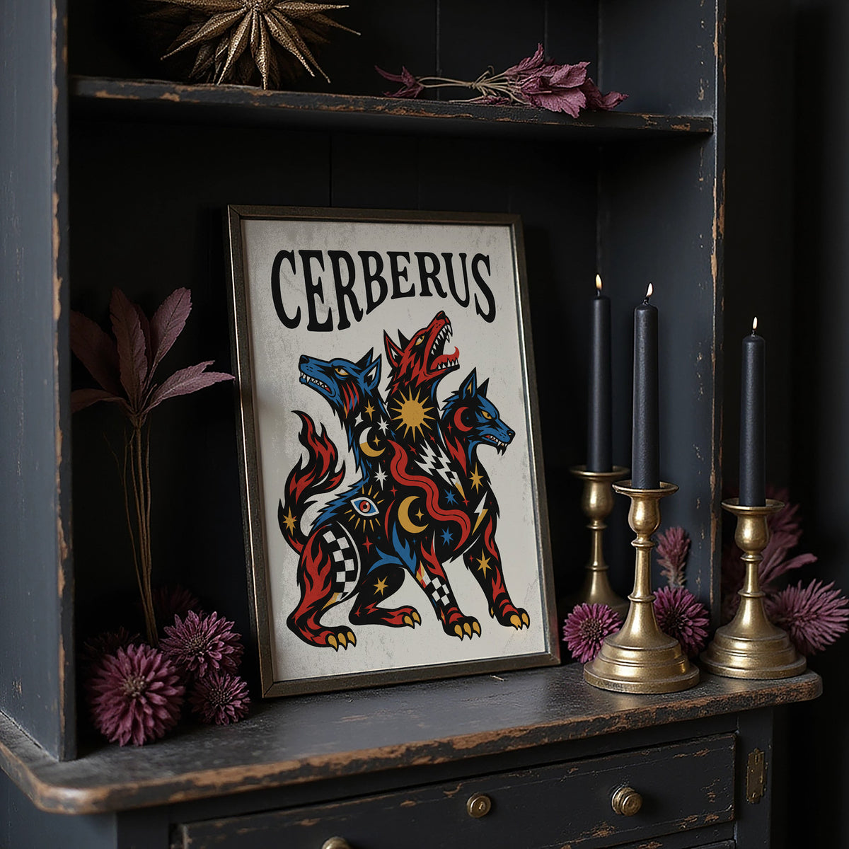 The Hound at the Threshold, a hound with several heads in red, blue and gold, with dark outlines and star and moon marks, in a bold flash sheet style with a black lettered name across the top, in a dark frame on a black shelf beside brass candles and dried leaves