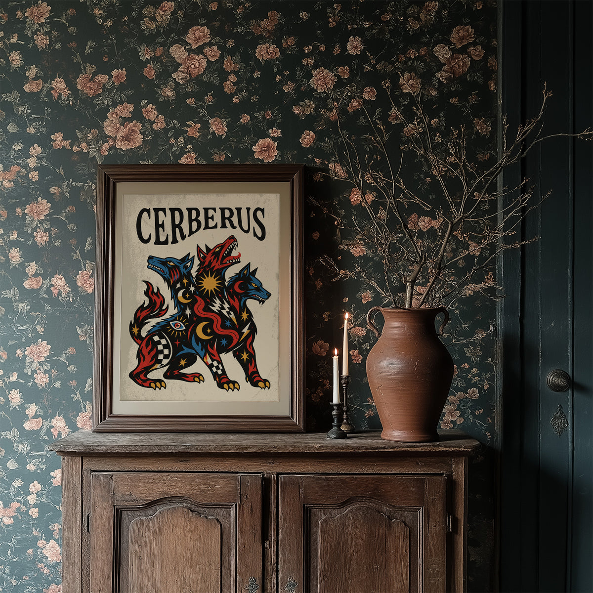 The Hound at the Threshold, a hound with several heads in red, blue and gold, with dark outlines and star and moon marks, in a bold flash sheet style with a black lettered name across the top, in a dark wood frame on a floral wallpaper wall above a rustic cabinet and a vase