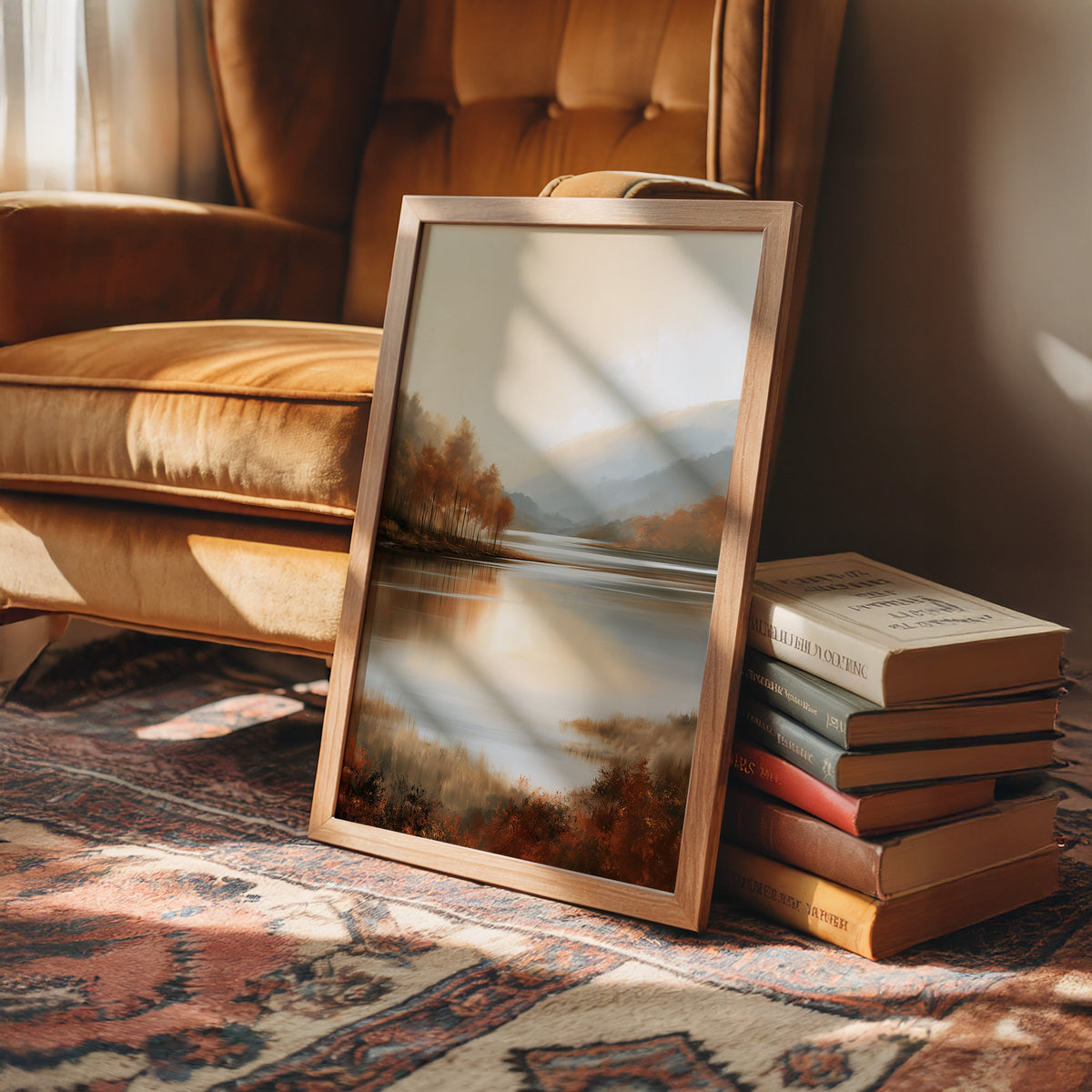 The Lake Remembers Autumn, an autumn lake at dusk with a line of turning trees on the near shore and pale mountains beyond, the water mirroring the sky in soft blue and amber, in a wood frame on a rug beside a leather armchair and a stack of books