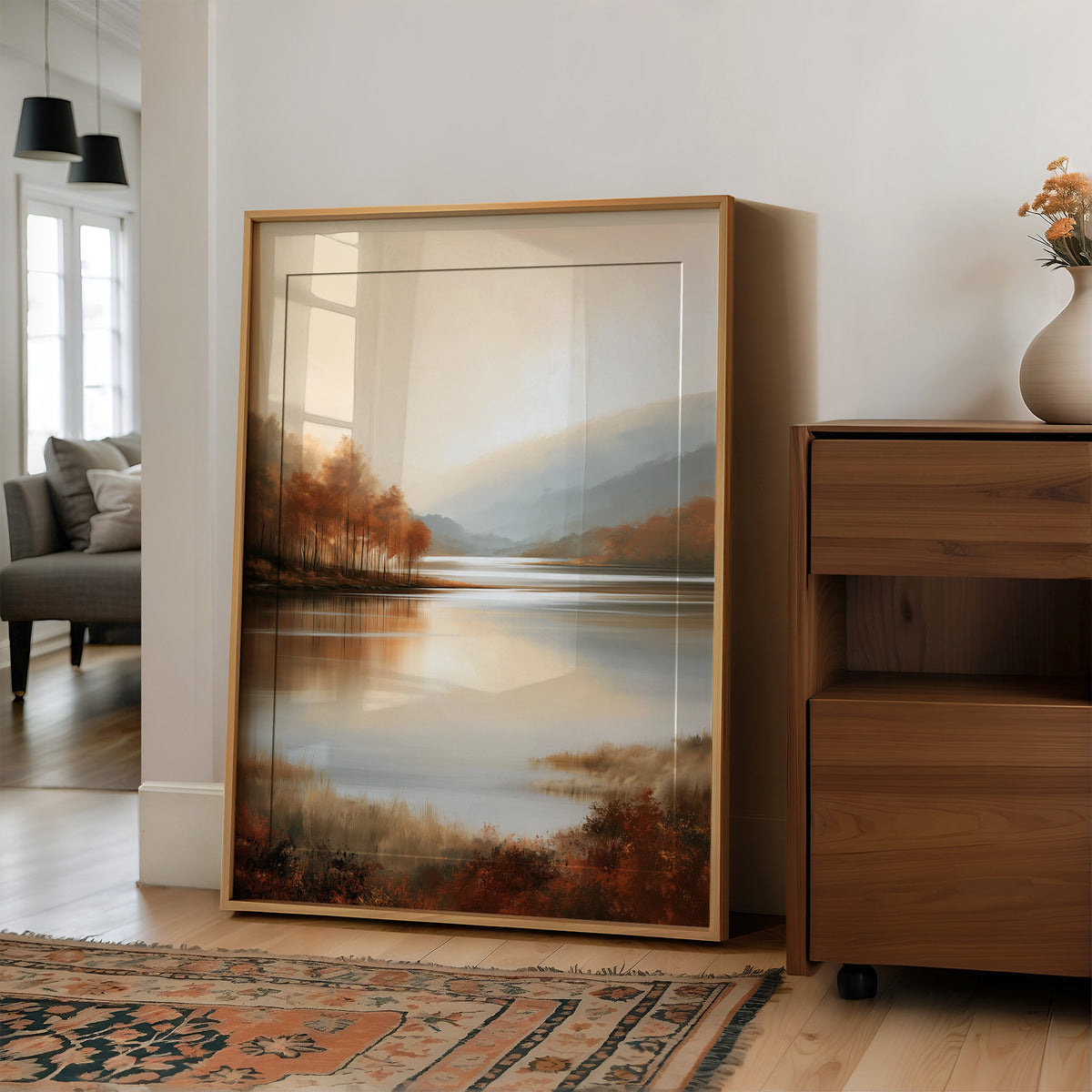 The Lake Remembers Autumn, an autumn lake at dusk with a line of turning trees on the near shore and pale mountains beyond, the water mirroring the sky in soft blue and amber, in a wood frame on a wall beside a walnut cabinet and an open doorway