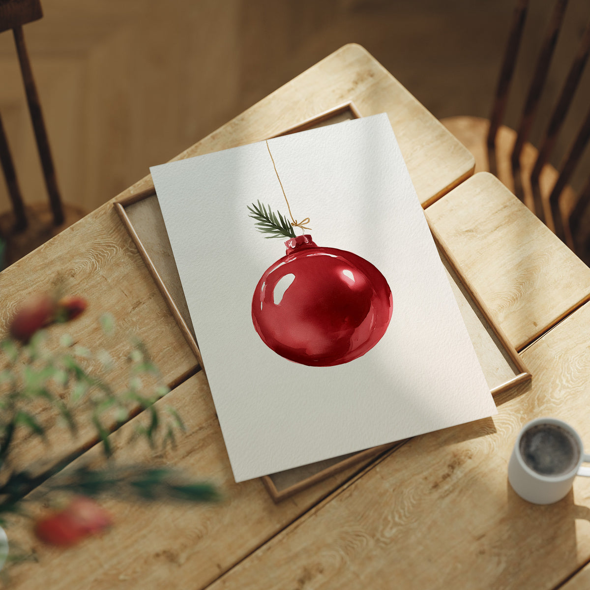 The Last Bauble on the Branch, a single red glass ornament hanging from a thin cord with a sprig of pine tip, on a cream ground, lying flat on a light wood table beside a coffee cup and red berries