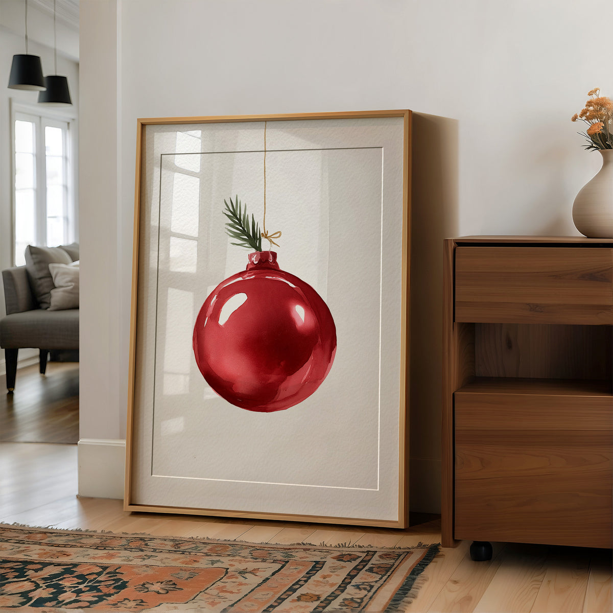 The Last Bauble on the Branch, a single red glass ornament hanging from a thin cord with a sprig of pine tip, on a cream ground, in a wood frame on a wall beside a walnut cabinet and an open doorway