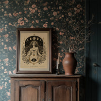 The Moon Woven Into Her Hair, a seated woman in quiet meditation crowned with a ring of crescent moons and surrounded by fern fronds, drawn in cream, charcoal and sepia with a deep brown border and small text at the base, in a dark wood frame on a floral wallpaper wall above a rustic cabinet and a vase