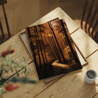The Path Bends Toward Home, an autumn forest path winding between tall dark trunks, lit by a shaft of golden light across the leaves, painted in rust, amber and deep brown, lying flat on a light wood table beside a coffee cup and red berries