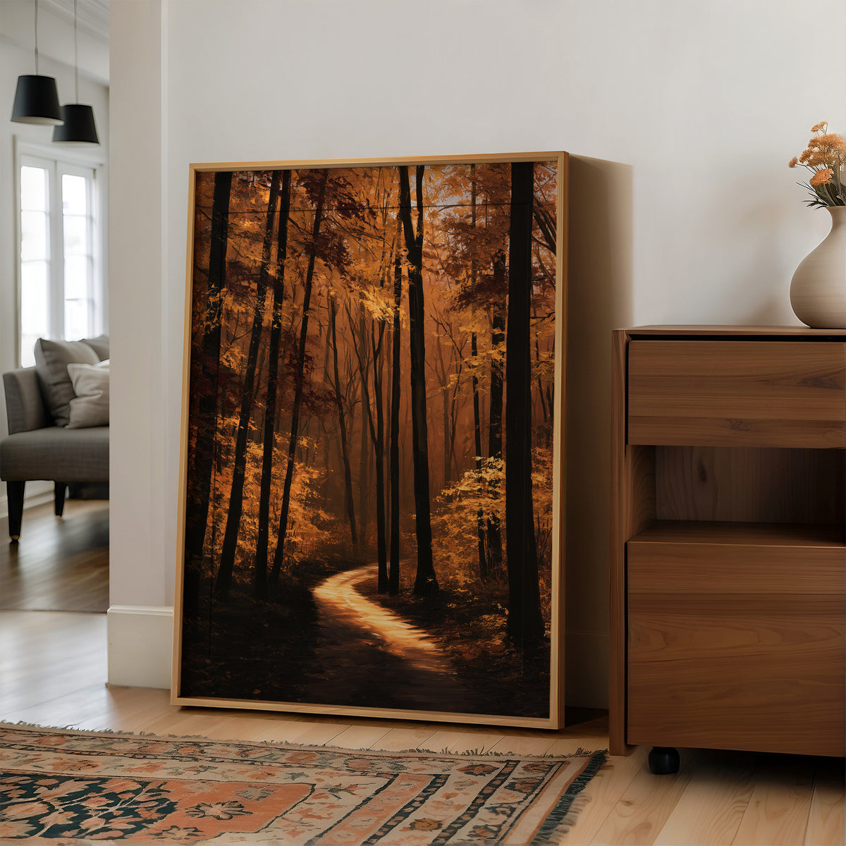 The Path Bends Toward Home, an autumn forest path winding between tall dark trunks, lit by a shaft of golden light across the leaves, painted in rust, amber and deep brown, in a wood frame on a wall beside a walnut cabinet and an open doorway