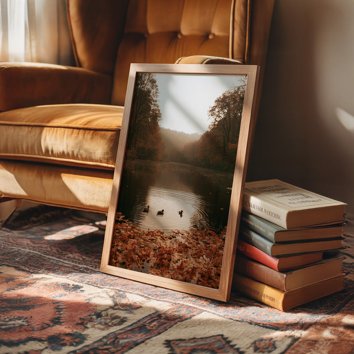 The Pond Keeps Its Quiet Company, two ducks on a still misty lake among rust trees, a bank of fallen autumn leaves in the foreground, painted in rust, forest green and soft gold, in a wood frame on a rug beside a leather armchair and a stack of books