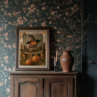The Pumpkin Keeps Its Hat On, a grinning jack o lantern wearing a witch hat on a weathered sign that reads happy halloween, with pumpkins and cornstalks, painted in burnt orange, black and aged cream, in a dark frame on a wooden dresser against floral wallpaper