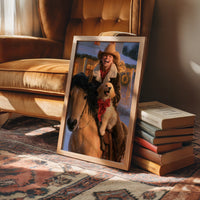 The Rider and the Golden Companion, a rider in a wide brimmed hat on a brown horse with a golden dog at the saddle, painted in saddle brown, dusky blue and warm gold, in a wood frame on a rug beside a leather armchair and a stack of books