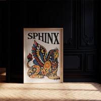 The Sphinx Keeps Her Riddle, a winged sphinx seated with folded wings in gold, red and blue, painted in bold black outlines on a cream paper ground beneath bold black lettering, in a traditional tattoo flash style, in a dark frame on a black paneled wall above a sunlit herringbone floor