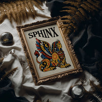 The Sphinx Keeps Her Riddle, a winged sphinx seated with folded wings in gold, red and blue, painted in bold black outlines on a cream paper ground beneath bold black lettering, in a traditional tattoo flash style, in an ornate frame on silk cloth beside ferns and a brass compass