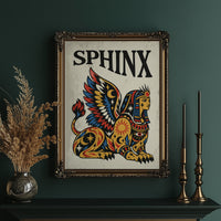 The Sphinx Keeps Her Riddle, a winged sphinx seated with folded wings in gold, red and blue, painted in bold black outlines on a cream paper ground beneath bold black lettering, in a traditional tattoo flash style, in an ornate gold frame on a deep green wall beside tall brass candlesticks