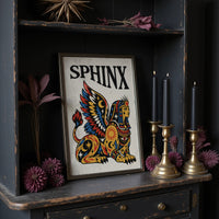 The Sphinx Keeps Her Riddle, a winged sphinx seated with folded wings in gold, red and blue, painted in bold black outlines on a cream paper ground beneath bold black lettering, in a traditional tattoo flash style, in a dark frame on a black shelf beside brass candles and dried leaves