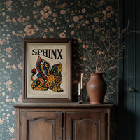The Sphinx Keeps Her Riddle, a winged sphinx seated with folded wings in gold, red and blue, painted in bold black outlines on a cream paper ground beneath bold black lettering, in a traditional tattoo flash style, in a dark wood frame on a floral wallpaper wall above a rustic cabinet and a vase