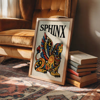 The Sphinx Keeps Her Riddle, a winged sphinx seated with folded wings in gold, red and blue, painted in bold black outlines on a cream paper ground beneath bold black lettering, in a traditional tattoo flash style, in a wood frame on a leather armchair beside a stack of books