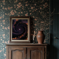The Spiral Remembers Everything, a violet and black galaxy turning around a bright core, edged with molten orange dust, in a dark frame on a wooden dresser against floral wallpaper