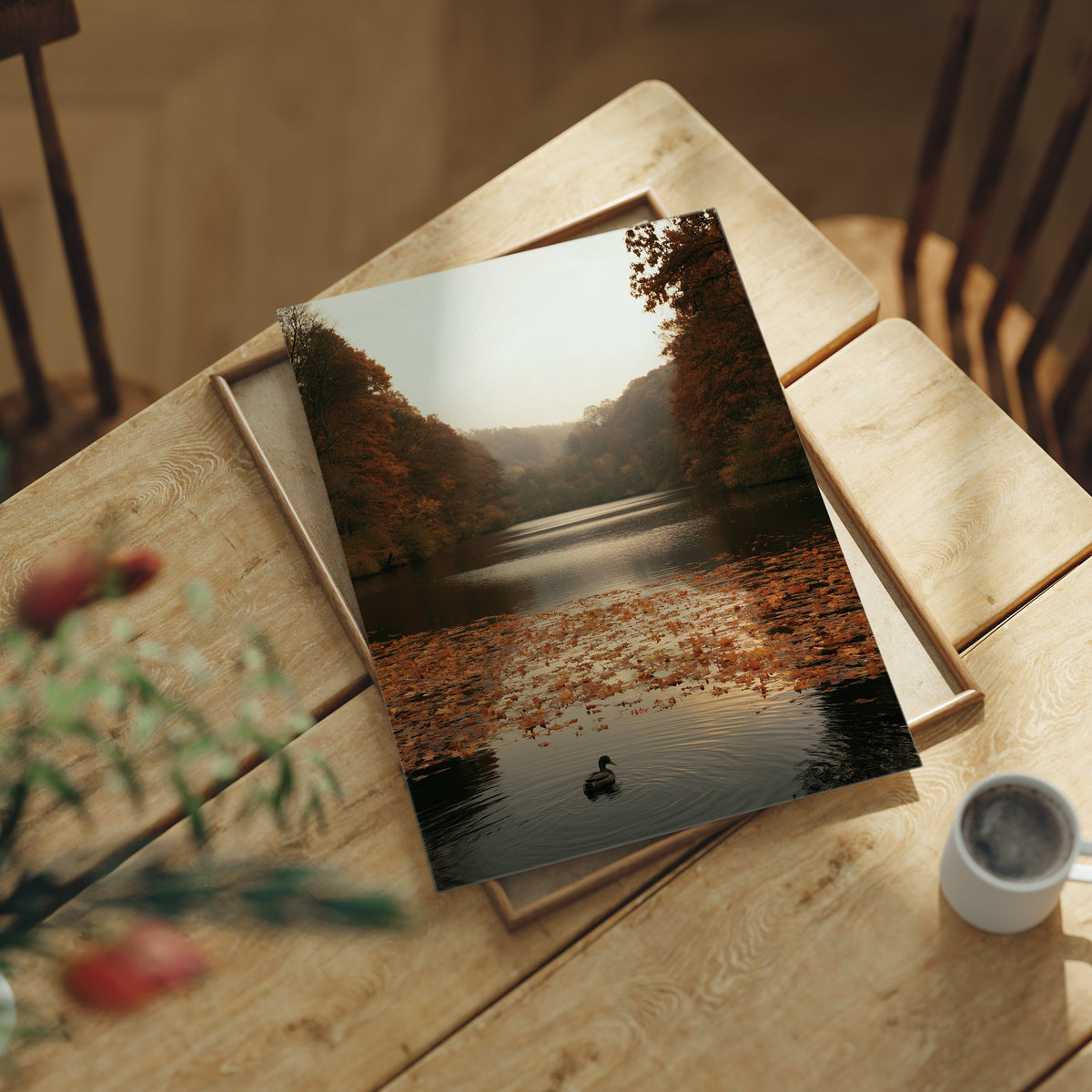 The Swan Waits on the Lake, a lone swan drifting on still water under turning autumn trees, with fallen leaves on the shore and a hazy golden light, painted in rust, amber and soft slate, lying flat on a light wood table beside a coffee cup and red berries