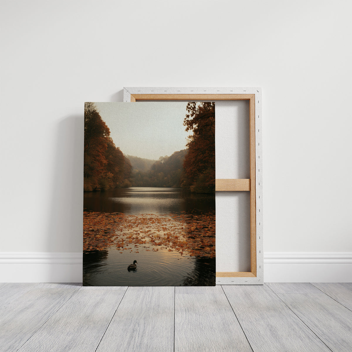 The Swan Waits on the Lake, a lone swan drifting on still water under turning autumn trees, with fallen leaves on the shore and a hazy golden light, painted in rust, amber and soft slate, on a gallery canvas with wrapped edges leaning against a white wall