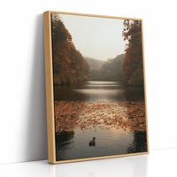 The Swan Waits on the Lake, a lone swan drifting on still water under turning autumn trees, with fallen leaves on the shore and a hazy golden light, painted in rust, amber and soft slate, as a framed canvas in a natural wood frame