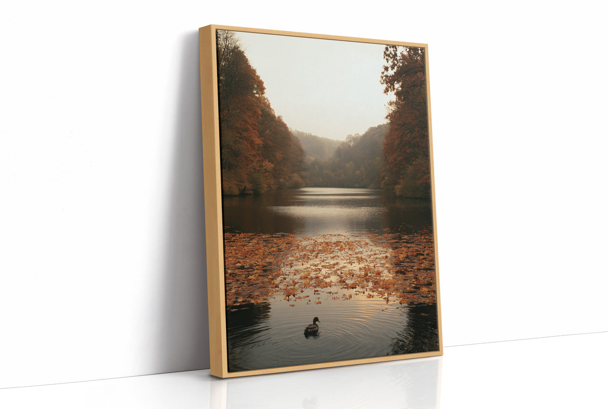 The Swan Waits on the Lake, a lone swan drifting on still water under turning autumn trees, with fallen leaves on the shore and a hazy golden light, painted in rust, amber and soft slate, as a framed canvas in a natural wood frame