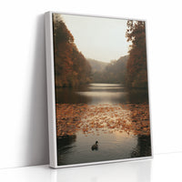 The Swan Waits on the Lake, a lone swan drifting on still water under turning autumn trees, with fallen leaves on the shore and a hazy golden light, painted in rust, amber and soft slate, as a framed canvas in a white frame