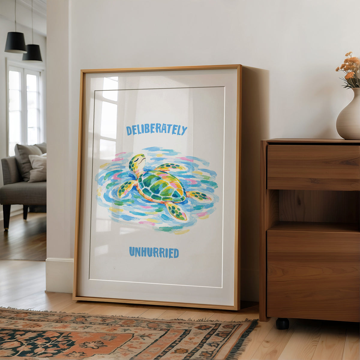 The Turtle Keeps Its Own Time, a sea turtle drifting over pale blue and green watercolor waves, with two lines of bold blue lettering above and below it on a white ground, in a soft watercolor style, in a wood frame on a wall beside a walnut cabinet and an open doorway
