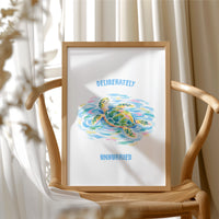 The Turtle Keeps Its Own Time, a sea turtle drifting over pale blue and green watercolor waves, with two lines of bold blue lettering above and below it on a white ground, in a soft watercolor style, in a wood frame beside a wicker chair by a sunny window