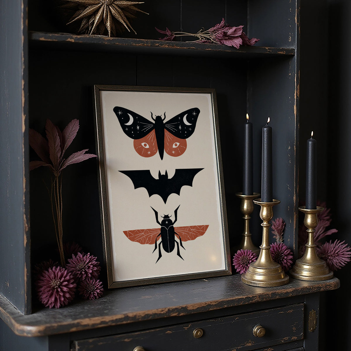 Three Night Visitors in a Row, a moth with crescent moon spots, a black bat in flight and a beetle with rust wings stacked in a column on a cream ground, painted in black, rust and cream, in a dark frame on a black shelf beside brass candles and dried leaves