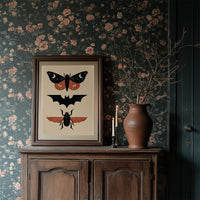 Three Night Visitors in a Row, a moth with crescent moon spots, a black bat in flight and a beetle with rust wings stacked in a column on a cream ground, painted in black, rust and cream, in a dark wood frame on a floral wallpaper wall above a rustic cabinet and a vase