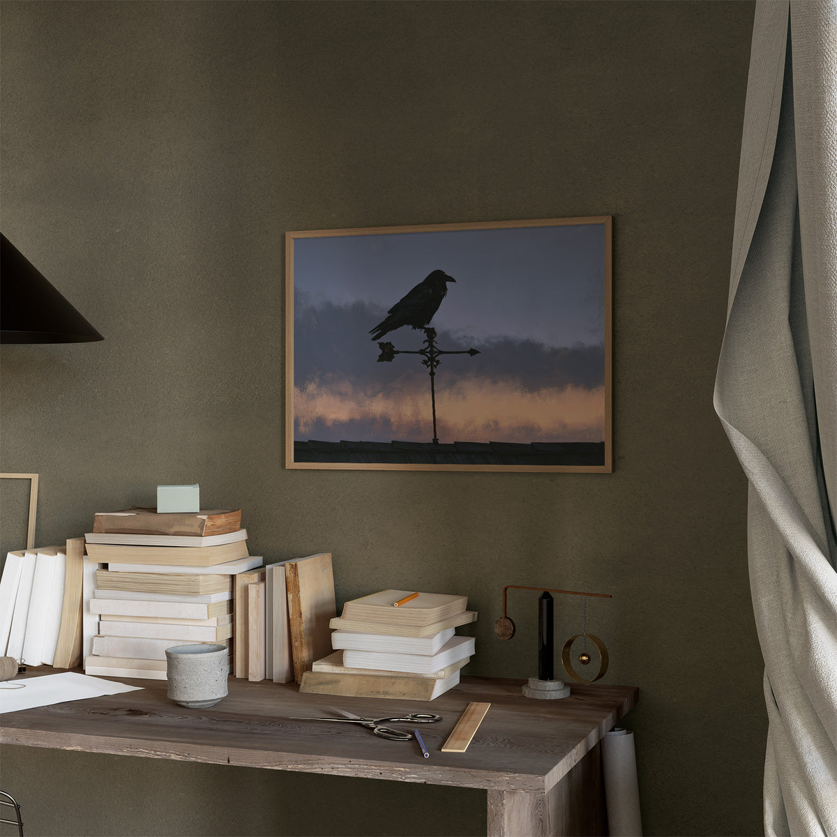 Watcher on the Weathervane, a black raven perched on a weathervane against a dusk sky banded in dusky blue and rose, in a thin wood frame on a moss green wall above a desk stacked with books