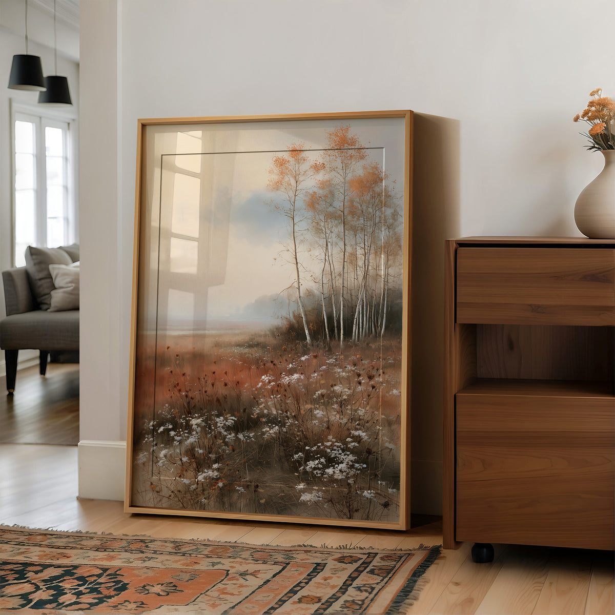 Where the Birches Lean Toward Dusk, a stand of pale birch trees with a few amber leaves in a misty meadow of rust grass and wildflowers under a soft blue sky, painted in dusty blue, rust and cream, in a wood frame on a wall beside a walnut cabinet and an open doorway