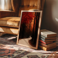 Where the Crimson Path Bends, a winding forest path through tall dark trunks under a blaze of red canopy, painted in deep crimson, russet and warm shadow, in a wood frame on a rug beside a leather armchair and a stack of books
