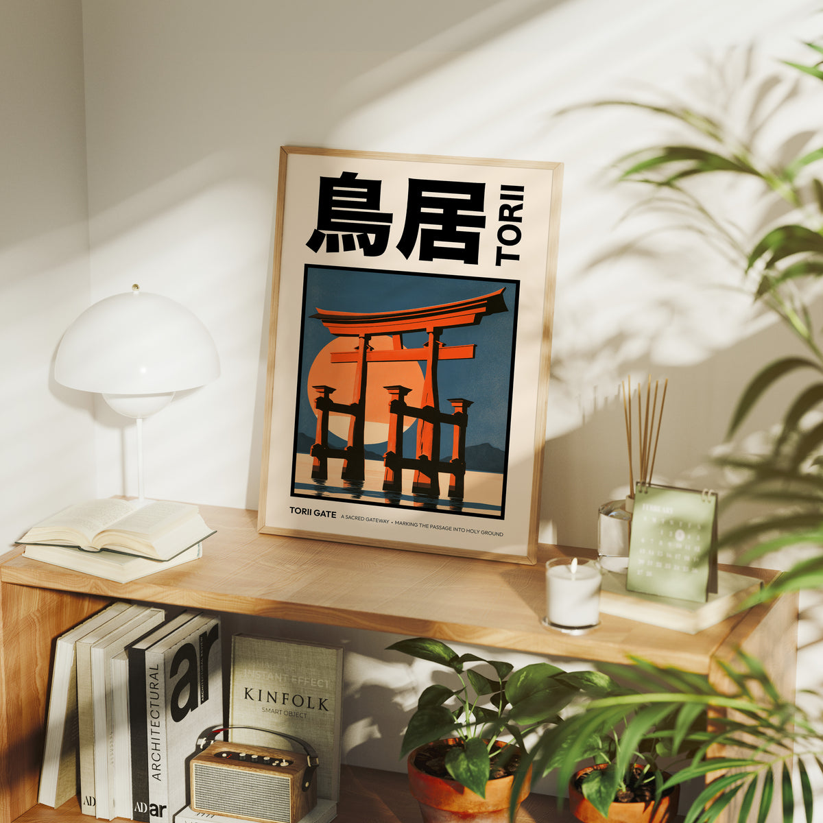 Where the Tide Meets the Posts, a vermilion torii shrine gate standing in shallow teal water beneath a bright orange sun, in a flat Japanese travel poster style with bold black lettering, painted in vermilion, teal and cream, in a wood frame on a console table beside a white lamp and stacked books
