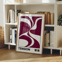 Where Two Curves Almost Touch, two maroon curved forms on a cream ground that lean toward each other and almost meet, with the word TENSION in black capitals and a small Japanese word beside it, in a wood frame on a white bookshelf beside stacked books and a small basket