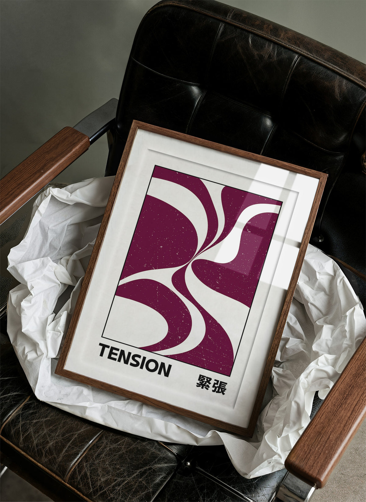 Where Two Curves Almost Touch, two maroon curved forms on a cream ground that lean toward each other and almost meet, with the word TENSION in black capitals and a small Japanese word beside it, in a wood frame on a black leather armchair beside crumpled white tissue